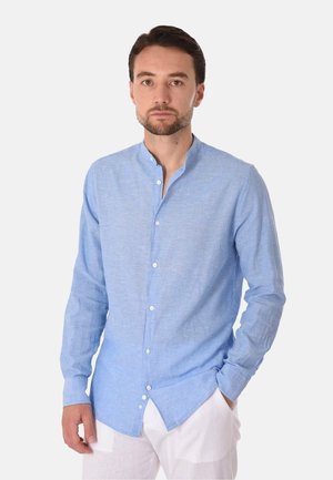Uomo con capelli corti castani, che indossa una camicia azzurro chiaro a maniche lunghe con bottoni e pantaloni bianchi, in piedi con una mano in tasca.