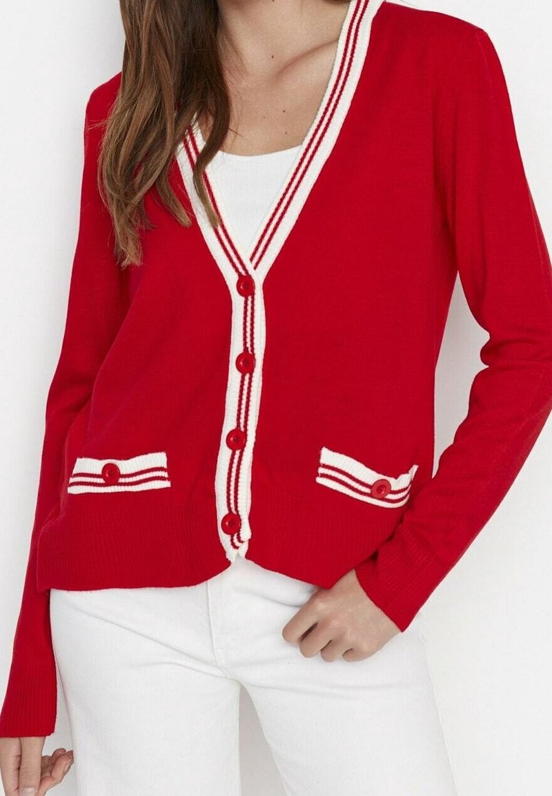Mujer vistiendo un cárdigan rojo con ribetes y botones blancos sobre una blusa blanca y pantalones blancos, con la mano descansando en la cadera.