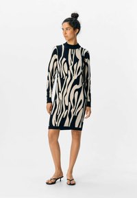 Donna con capelli scuri raccolti in uno chignon, che indossa un abito lungo a maniche lunghe con una fantasia abstract in nero e beige e sandali con tacco neri, in piedi e che guarda di lato.