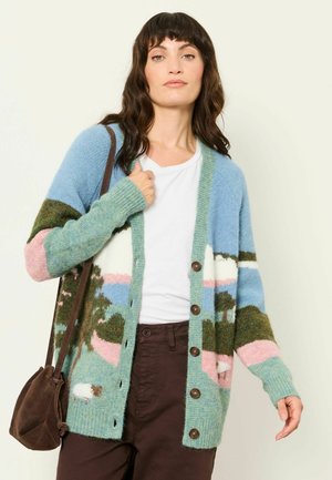 Woman wearing a colorful patterned cardigan with trees and sheep, white shirt, brown pants, and carrying a brown shoulder bag.