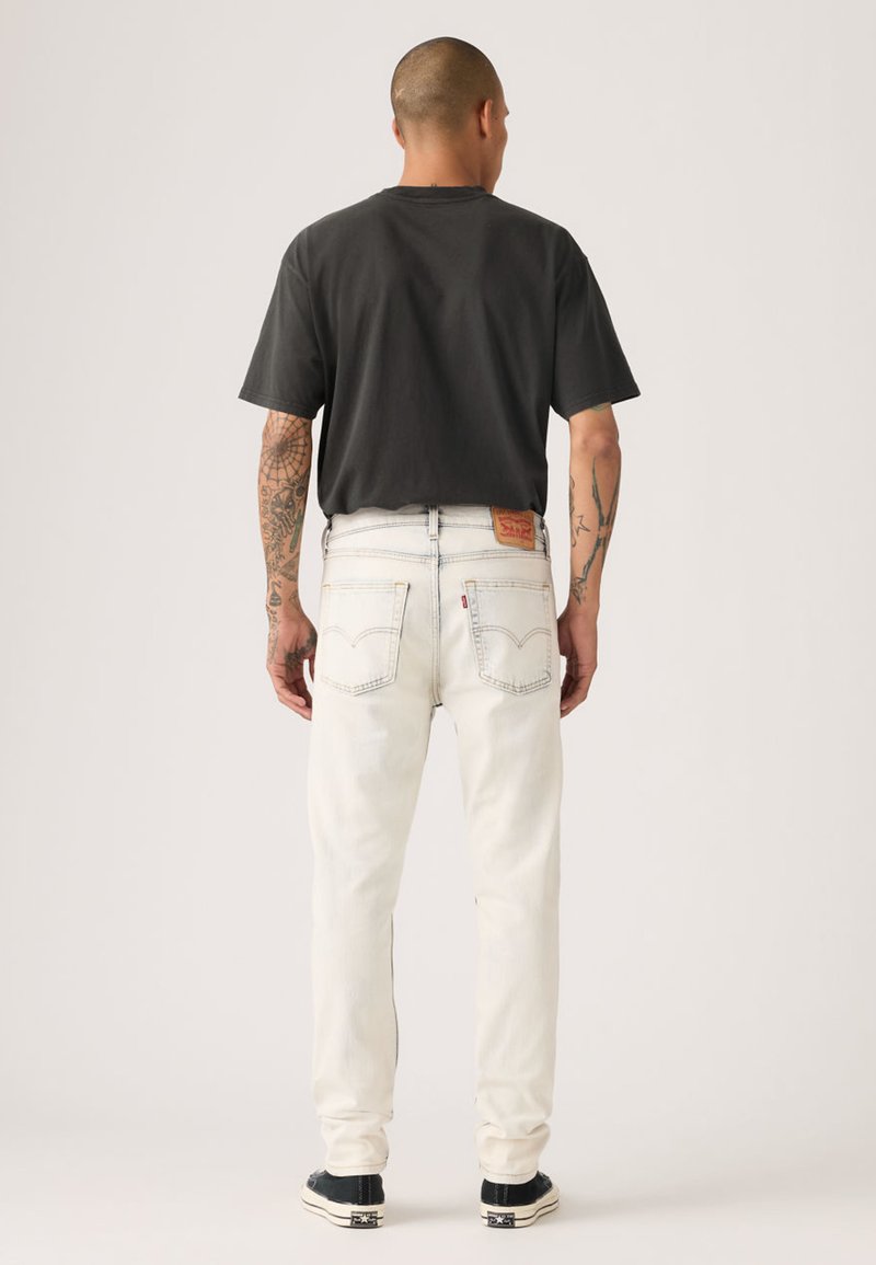 Jean en denim blanc à coupe droite, montrant des poches arrière et des coutures contrastantes. Associé à un t-shirt noir et des baskets.