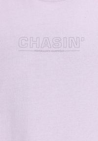 Šviesiai violetinė medvilnės mišinio marškinėlių viršus, su tekstūruotu atspaudu „CHASIN'“ tamsiai pilkos spalvos, po kuriuo yra užrašas „PERFORMANCE JEANSWEAR.“