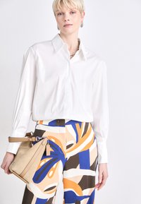 White button-up shirt with a collar, paired with patterned trousers featuring orange, blue, and brown organic shapes. Carrying a beige handbag.