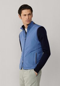 Muž v modré vestičce na zip bez rukávů přes černé tričko s dlouhým rukávem, s rukama v kapsách světle šedých kalhot, dívající se stranou.