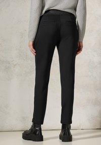 Black, fitted trousers with a smooth texture, featuring a solid waistband and two back pockets. Styled with a grey sweater and ankle boots.