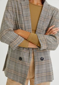 Kostkovaný dvoubarevný blejzr v šedé, hnědé a černé barvě. Má uvolněný střih a ohrnuté rukávy. Pod ním je hnědý žebrovaný top.