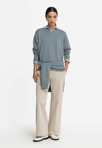 Hellblaues V-Neck-Sweater mit gerippten Bündchen, getragen über einem gestreiften Hemd mit Kragen; kombiniert mit cremefarbenen weiten Hosen und weißen Schuhen.