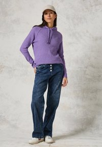 Lila gestrickter Pullover mit Kapuze, kombiniert mit weiter geschnittenen, dunkelblauen Jeans aus Denim mit Knopfdetails auf der Vorderseite. Weiße Sneakers dazu getragen.