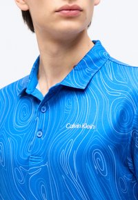 Άτομο που φοράει μπλε πόλο Calvin Klein με κυματιστά μοτίβα γραμμών, γιακά και δύο ορατά κουμπιά, πάνω σε λευκό φόντο.