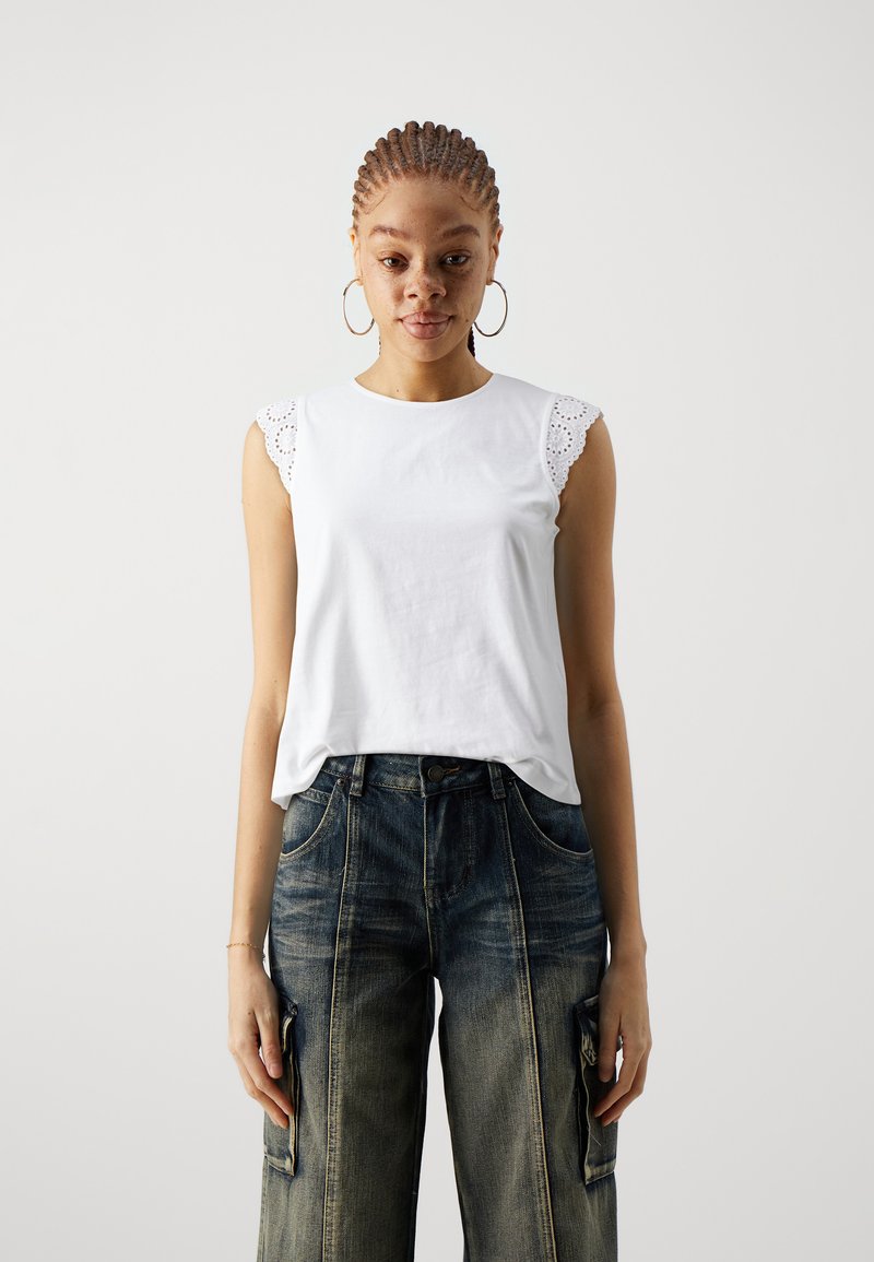 Top de algodón blanco sin mangas con ribete de ojales en las mangas, combinado con pantalones cargo de mezclilla azul oscuro que cuentan con bolsillos laterales y un corte relajado.