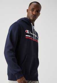 Jūras zila kapuce ar svārku kokvilnas sajaukumā, ar baltu saiti, priekšējo kabatu un "Champion New York" logotipu ar sarkaniem un baltiem svītrām.