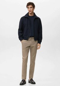 Giacca navy con zip sopra una maglia scura, abbinata a pantaloni beige a gamba stretta e mocassini neri. Tessuto liscio con design minimalista.