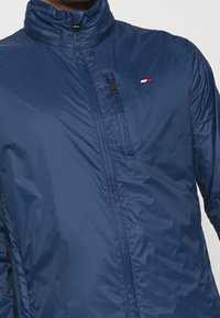 Chaqueta ligera de color azul marino con un alto cuello, cierre de cremallera y un pequeño logo. Presenta una textura suave y brillante con un diseño minimalista.