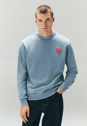 Mladý muž s krátkymi vlasmi, oblečený v svetlomodrej mikine s nášivkou červeného srdca a tmavých džínsoch, stojaci na jednoduchom pozadí.