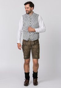 Grau gemusterte Weste über einem weißen Hemd, kombiniert mit braunen Lederhosen mit floraler Stickerei und dekorativen Knöpfen. Braune Stiefel.