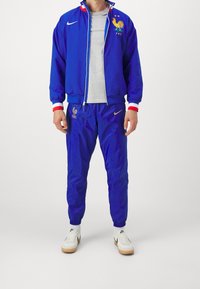Blå track jakke og bukser lavet af letvægtsmateriale, med et hanemblem, Nike-logo og røde og hvide detaljer på manchetten.