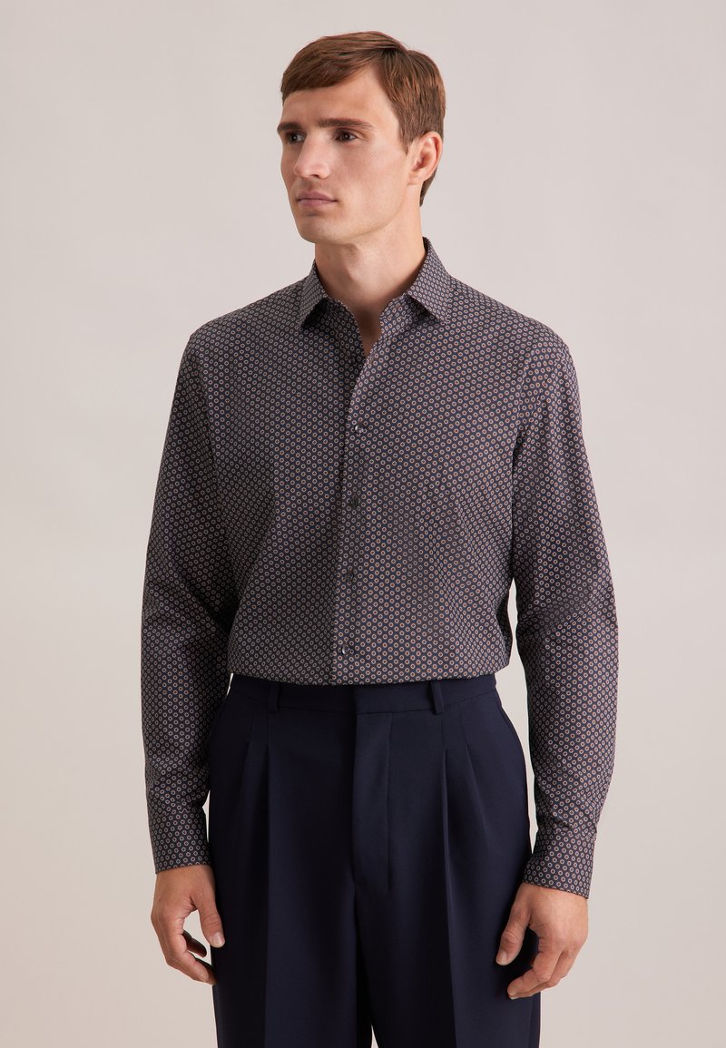 Camicia botton-down blu navy con un motivo geometrico in rosso, bianco e azzurro chiaro. Presenta un colletto e maniche lunghe; abbinata a pantaloni scuri.