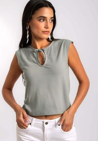 Top sin mangas de color verde claro con escote redondo y detalle de ojal, que presenta pequeños adornos a lo largo del dobladillo y una textura de tela suave.