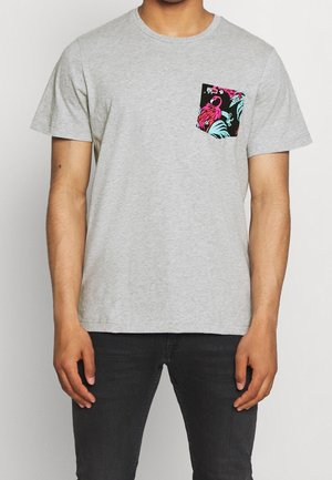 Mężczyzna w jasno szarej koszulce z krótkim rękawem i małą czarną kieszonką z wzorem różowego flaminga i turkusowych liści.