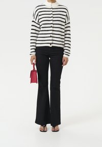 Musta-valkoraidallinen neulottu neulejakku, jossa on napit, yhdistetty tummiin liehukkeisiin farkkuihin. Oikeassa kädessä pidetään pientä punaista käsilaukkua.