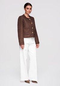 Žena stojící, oblečená v hnědém texturovaném zapínacím saku, bílých kalhotách s širokými nohavicemi a hnědých lodičkách s špičatou špičkou na jednobarevném pozadí.