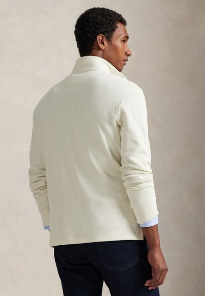 Άνδρας που φοράει κρεμ πουκάμισο μακρύ μανίκι με ψηλό γιακά, σκούρα τζιν, στέκεται πλάγια μπροστά σε απλό ανοιχτό φόντο.