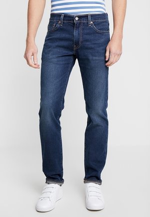 Jeans de mezclilla azul oscuro con corte recto, que cuentan con cinco bolsillos, pasadores para cinturón y costuras en contraste. Llevados con zapatillas blancas.