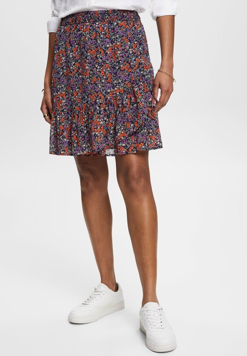 Femme portant une jupe courte à fleurs avec des fleurs orange et violettes, des baskets blanches et une chemise blanche à manches longues avec les manches retroussées.