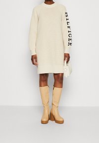Abito in maglia crema con texture a coste e maniche oversized; testo nero "HILFIGER" su un braccio. Abbinato a stivali platform color cammello al ginocchio.
