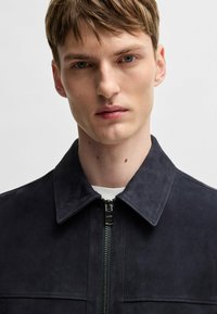 Navy suede jacket with a pointed collar, front zipper closure, and contrast stitching. Smooth texture with structured design elements.