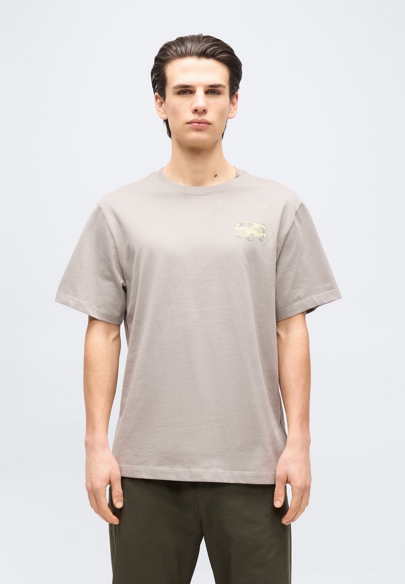 Young man with slicked-back hair wearing a light gray T-shirt with small van graphic on chest, standing against plain background.