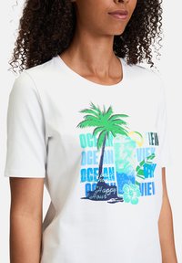 T-shirt blanc à manches courtes présentant un graphisme coloré d'un palmier, d'une boisson et du texte "Vue sur l'Océan" dans diverses nuances de bleu et de vert.