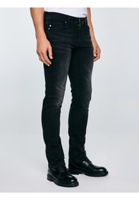 Férfi fekete, testhezálló farmerban és fekete bőr, fűzős cipőben, semleges háttér előtt állva.