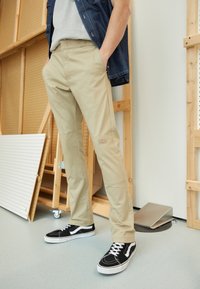 Beige pants with a straight cut, featuring knee patches and a logo. Worn with black sneakers with white accents. Background includes wood and tools.