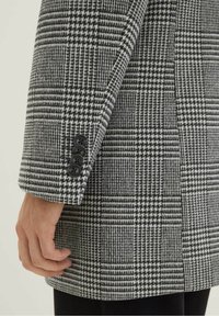 Szary i czarny płaszcz w kratę o dopasowanym kroju, teksturowanej tkaninie oraz czterech czarnych guzikach na mankiecie. Zbliżenie na rękaw i dłoń.
