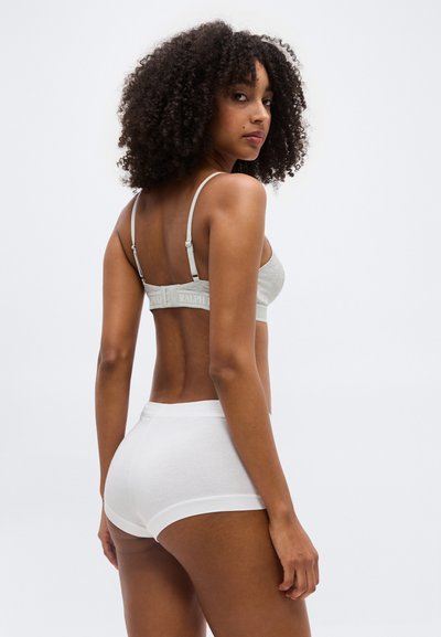 Γυναίκα με σγουρά μαλλιά που φοράει ανοιχτό γκρι σουτιέν και λευκά σορτς, γυρισμένη ελαφρώς για να δείξει την πλάτη της και κοιτάζοντας πάνω από τον ώμο της.