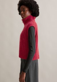 Fuchsia Strickweste mit hohem Kragen, mit weicher Textur und cropped Design, getragen über einem langärmeligen schwarzen Oberteil.