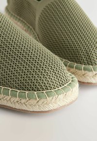 Espadrilles en tissu maille verte avec semelle en jute tressée et détails cousus sur un fond clair, en gros plan.