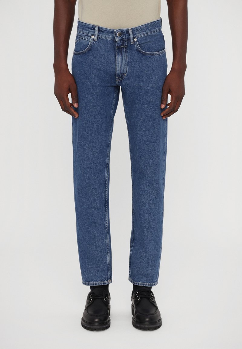 Pantalones vaqueros de mezclilla azul de pierna recta con un acabado desgastado, cinco bolsillos y cierre de botón, combinados con zapatos negros.