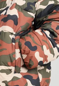 Transformuojantis pūkinė striukė kamufliažo stiliaus žalios, rudos, smėlio ir tamsiai mėlynos spalvos. Savybės: siūta medžiaga, per didelis kirpimas ir paslėptos kišenės.