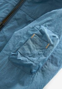 Blå tygjacka med en strukturerad yta, med en ficka med gula sömmar på överarmen. Dragkedjedetalj synlig.