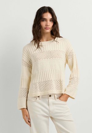 Donna con capelli scuri ondulati indossa un maglione di maglia crema lavorata e jeans chiari, in piedi con una mano in tasca su sfondo bianco.