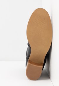 Chaussure en cuir noir avec une semelle en caoutchouc marron, présentant une finition lisse, une forme arrondie et une couture décorative près du talon.