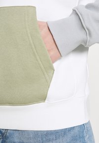 Huvtröja med en ficka dekorerad med en olivgrön panel, vit kropp och grå ärmslut. Slät textur med ribbad nederkant.