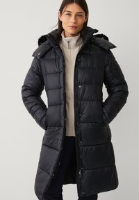 Sort puffer-jakke med aftagelig hætte, kviltet design, lynlås- og trykknaplukning samt sidelommer. Båret over en lys beige top.