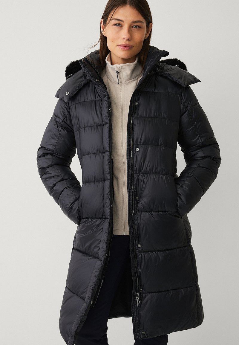 Sort puffer-jakke med aftagelig hætte, kviltet design, lynlås- og trykknaplukning samt sidelommer. Båret over en lys beige top.