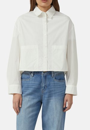 Vrouw die een witte cropped blouse met lange mouwen draagt, gecombineerd met high-waisted blauwe spijkerbroeken, staand tegen een effen achtergrond.