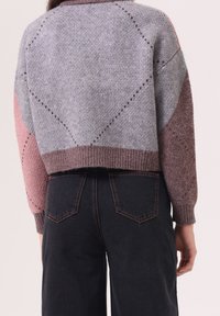 Pull en maille épaisse gris, rose et bordeaux avec un design court, des manches raglan et des motifs texturés. Jean foncé visible en dessous.