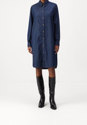 Persoon draagt een knielange jurk van donker denim met lange mouwen, knopen en een kraag, gecombineerd met zwarte lederen laarzen tot net onder de knie.