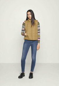 Colete acolchoado bege com gola alta, usado sobre uma camisa de mangas longas com riscas multicoloridas. Jeans ajustados azuis e sapatos pretos de atar completam o visual.